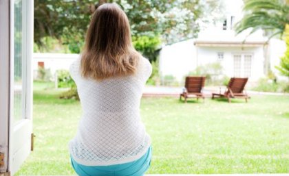 The findings suggest greenspace around a person's home can lead to more physical exercise, more Vitamin D and/or can mitigate the effects of air pollution. Image: Adobe. A woman sits at an open doorway with her back to the camera. She is facing a garden with a green lawn, trees and wooden chairs.